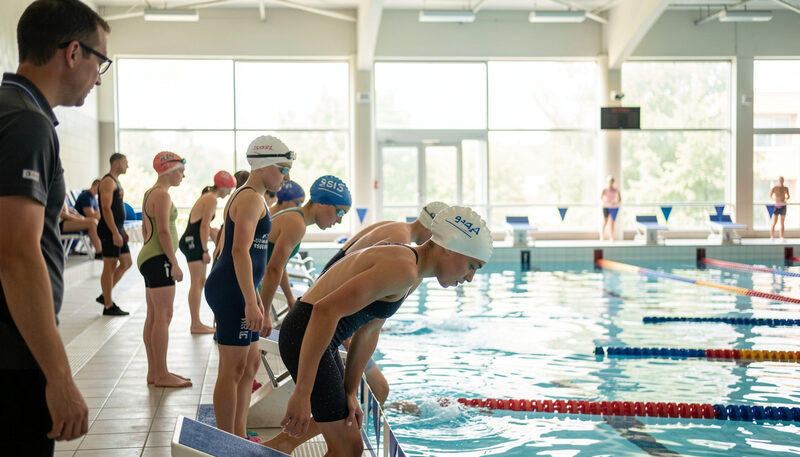 Young swimmers lined up on the starting blocks at an indoor pool as a race begins, a coach watching from the side.