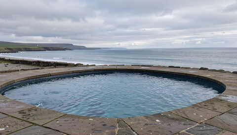 A coastal seawater pool on a stone promenade at high tide, cold blue water and a cloudy Scottish sky.